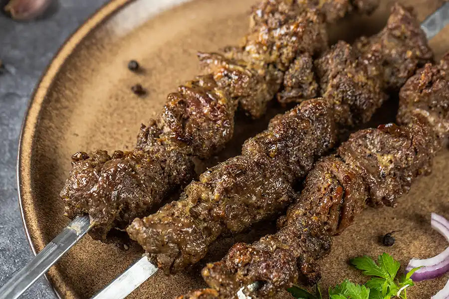 Lamm-Schaschlik Knoblauch-Pfeffer auf einem rustikalen Teller mit Petersilie, Pfefferkörnern und roten Zwiebeln angerichtet.