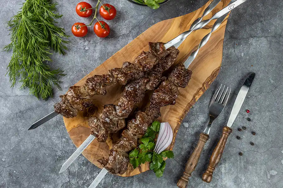 Lamm-Schaschlik Original serviert auf einem Holzbrett, angerichtet mit Petersilie und roten Zwiebeln. Dekoriert mit Pfefferkörnern, Cocktail-Tomaten, Kräutern und Besteck.