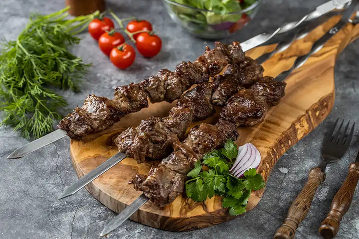 Lamm-Schaschlik Original serviert auf einem Holzbrett, angerichtet mit Petersilie und roten Zwiebeln. Dekoriert mit Pfefferkörnern, Cocktail-Tomaten, Kräutern und Besteck.