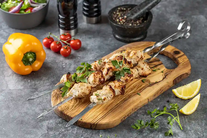 Goldbraun gegrilltes Poulet-Schaschlik Original auf einem Holzbrett, dekoriert mit Petersilie und Schnittlauch. Serviervorschlag mit Paprika, Cherrytomaten, Zitrone, Salat und Gewürzen.