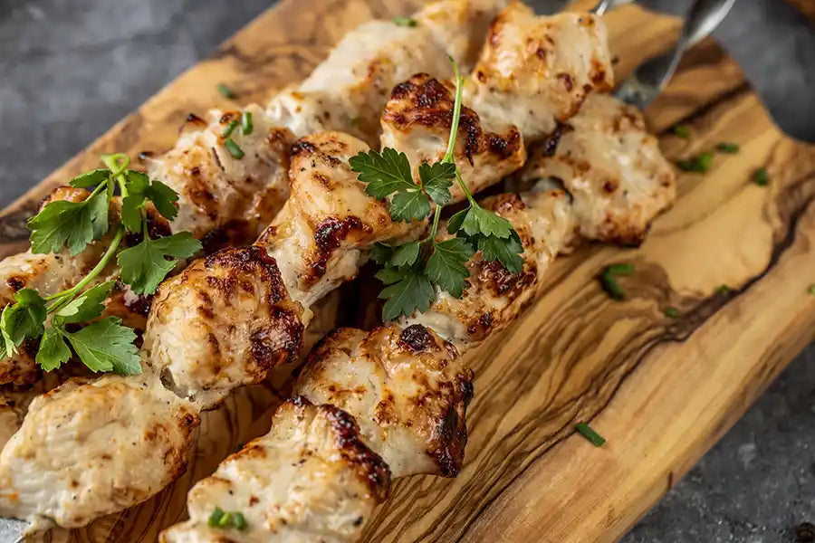 Nahaufnahme von goldbraun gegrilltem Poulet-Schaschlik auf Spießen, angerichtet auf einem Holzbrett und dekoriert mit Petersilie und Schnittlauch. Serviervorschlag.