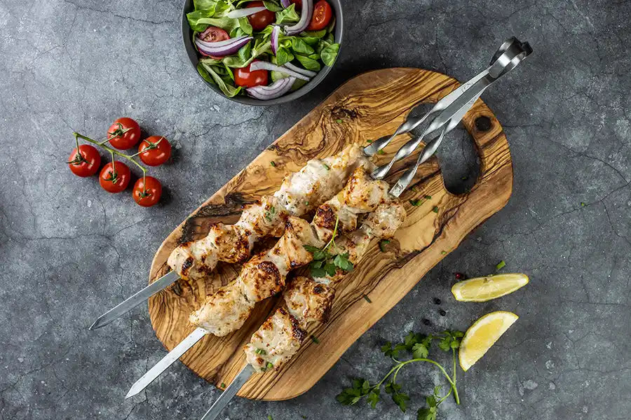 Goldbraun gegrilltes Poulet-Schaschlik Original auf einem Holzbrett, garniert mit Petersilie und Schnittlauch, serviert mit Cherrytomaten, Zitrone und Salat.