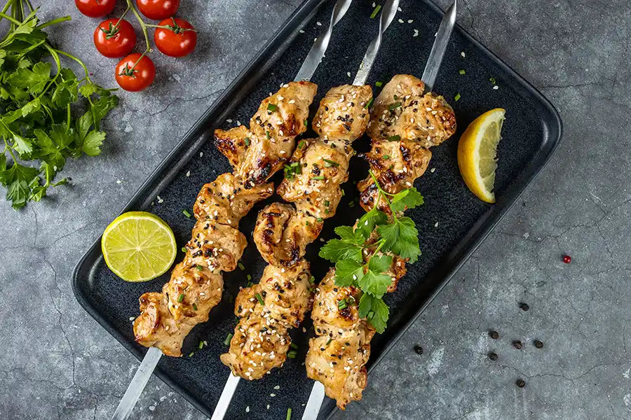 Poulet-Schaschlik Teriyaki goldbraun gegrillt, dekoriert mit Sesam, Schnittlauch, Gewürzen und Petersilie. Angerichtet auf einem Teller. Mit Cherry Tomaten,  Zitronen und Pfefferkörner.