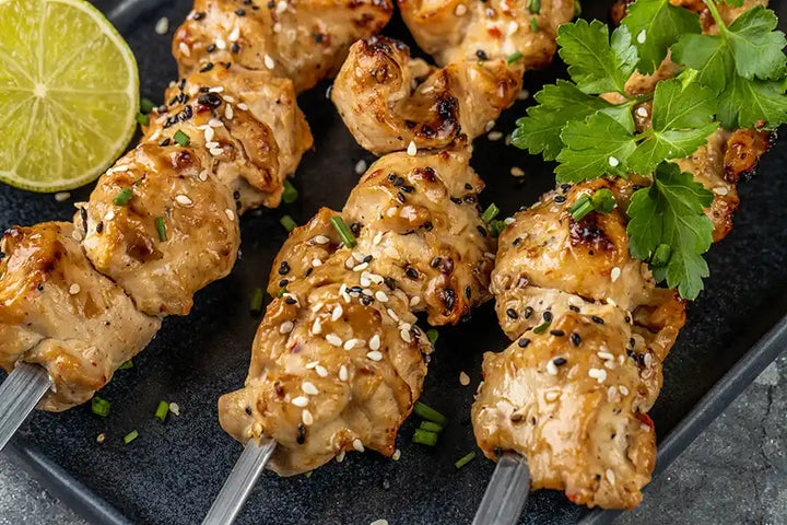 Poulet-Schaschlik Teriyaki goldbraun gegrillt in der Nahaufnahme, dekoriert mit Sesam, Schnittlauch, Gewürzen und Petersilie. Angerichtet auf einem Teller mit einer Zitronenscheibe.