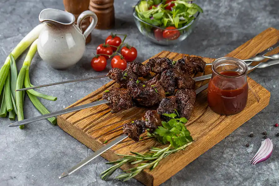 Rindfleisch-Schaschlik BBQ garniert mit Schnittlauch, Petersilie, Rosmarin und Sosse. Auf einem rustikalen Holzbrett angerichtet. Dekoriert mit Cherry-Tomaten, Salat, roten Zwiebeln, Pfefferkörnern und Frühlingszwiebeln.