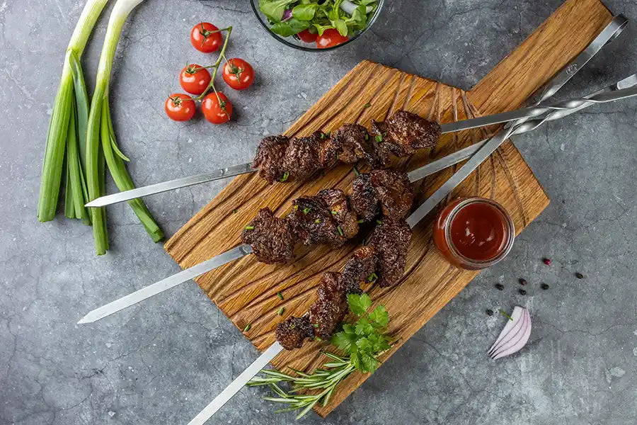 Rindfleisch-Schaschlik BBQ garniert mit Schnittlauch, Petersilie, Rosmarin und Sosse. Auf einem rustikalen Holzbrett angerichtet. Dekoriert mit Cherry-Tomaten, Salat, roten Zwiebeln, Pfefferkörnern und Frühlingszwiebeln.