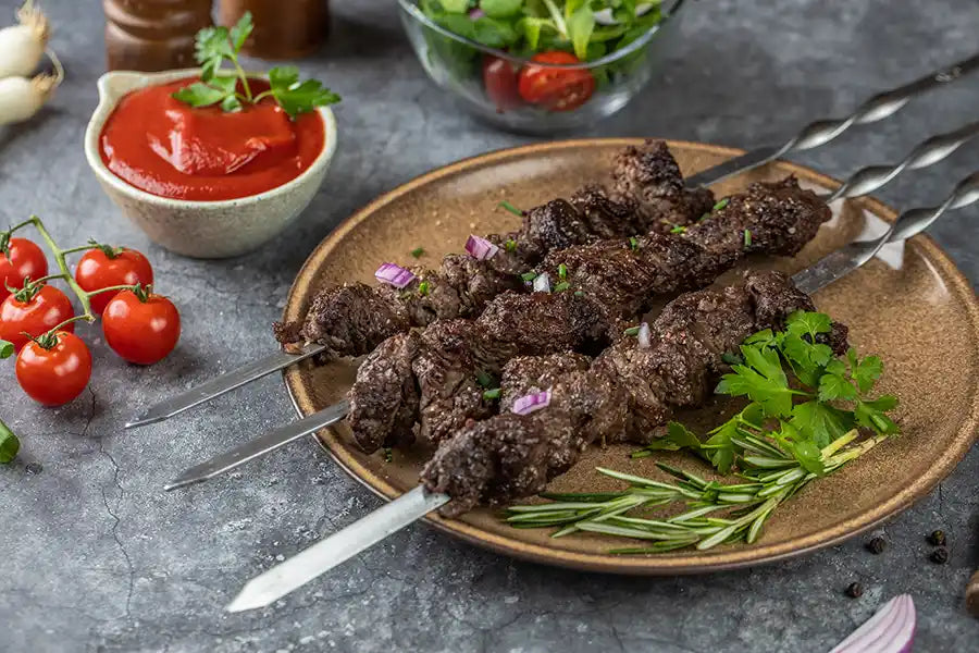 Rindfleisch Schaschlik nach Original Rezept garniert mit roten Zwiebeln, Schnittlauch, Petersilie und Rosmarin. Angerichtet auf einem Teller. Dekoriert mit Salat, Sosse, Cocktailtomaten und Pfefferkörnern.