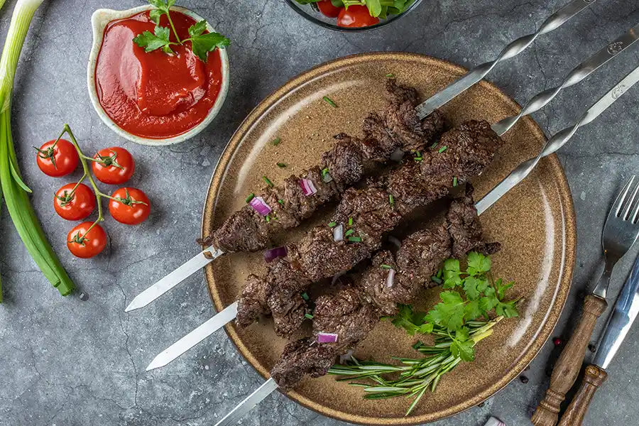 Rindfleisch Schaschlik nach Original Rezept garniert mit roten Zwiebeln, Schnittlauch, Petersilie und Rosmarin. Angerichtet auf einem Teller. Dekoriert mit Salat, Sosse, Cocktailtomaten, Pfefferkörnern und Besteck.