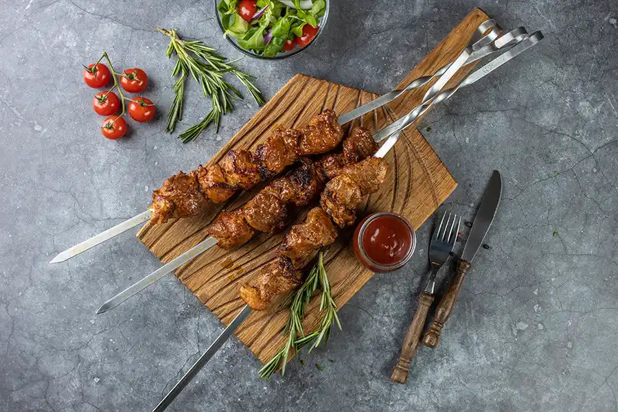 Schweine-Schaschlik BBQ angerichtet auf einem rustikalen Holzbrett mit Barbecue-Sosse und Rosmarin. Dekoriert mit Salat, Cocktail-Tomaten, Besteck und buntem Pfeffer.