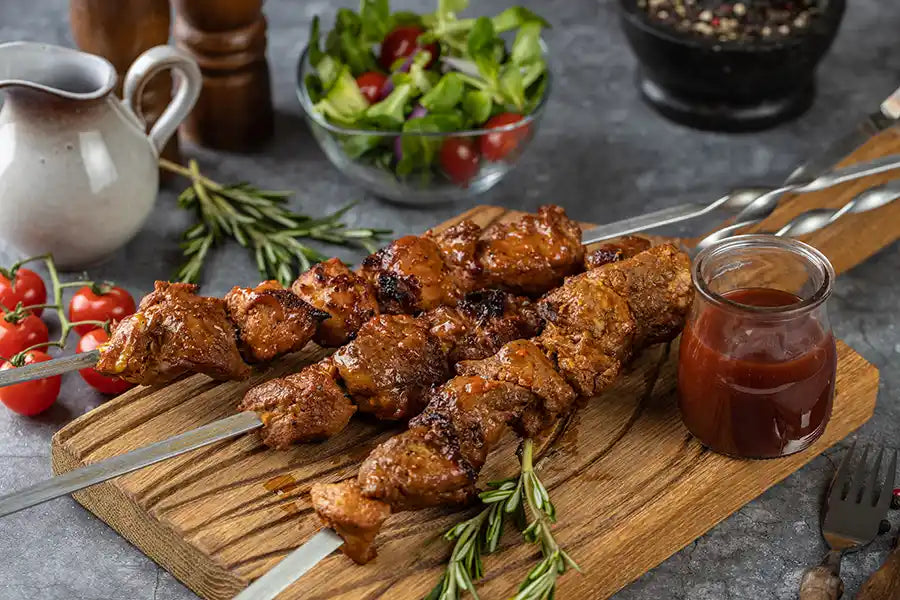 Schweine-Schaschlik BBQ angerichtet auf einem rustikalen Holzbrett mit Barbecue-Sosse und Rosmarin. Dekoriert mit Salat, Cocktail-Tomaten, Besteck, buntem Pfeffer und einer Sauciere.