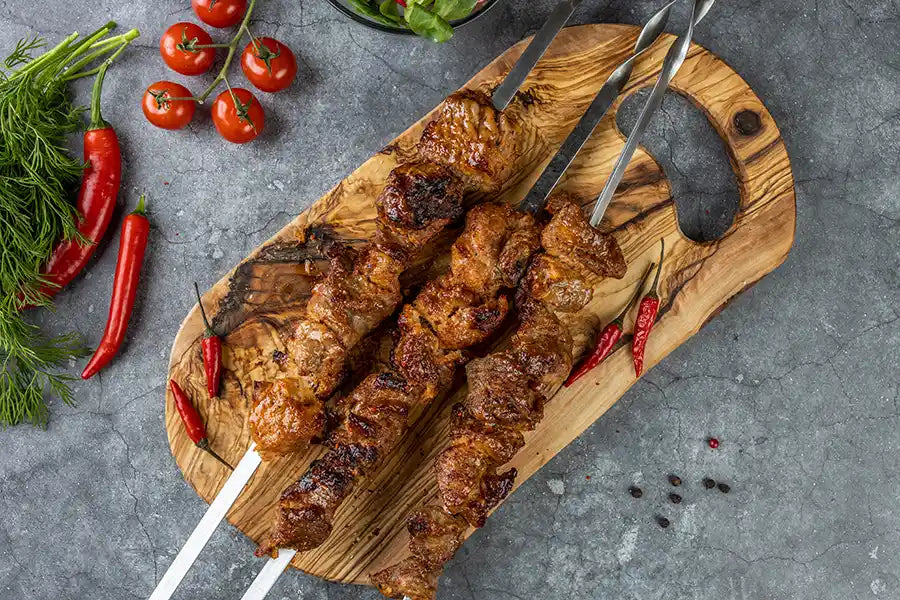 Schweine-Schaschlik Chili auf einem Holzbrett angerichtet mit Chilischoten. Dekoriert mit Cocktail-Tomaten, Salat und bunten Pfefferkörnern.