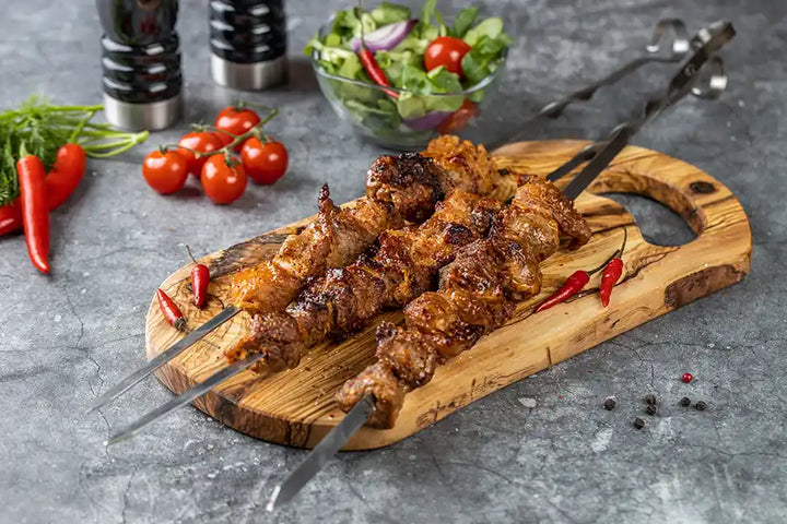 Schweine-Schaschlik Chili auf einem Holzbrett angerichtet mit Chilischoten. Dekoriert mit Cocktail-Tomaten, Salat und bunten Pfefferkörnern.