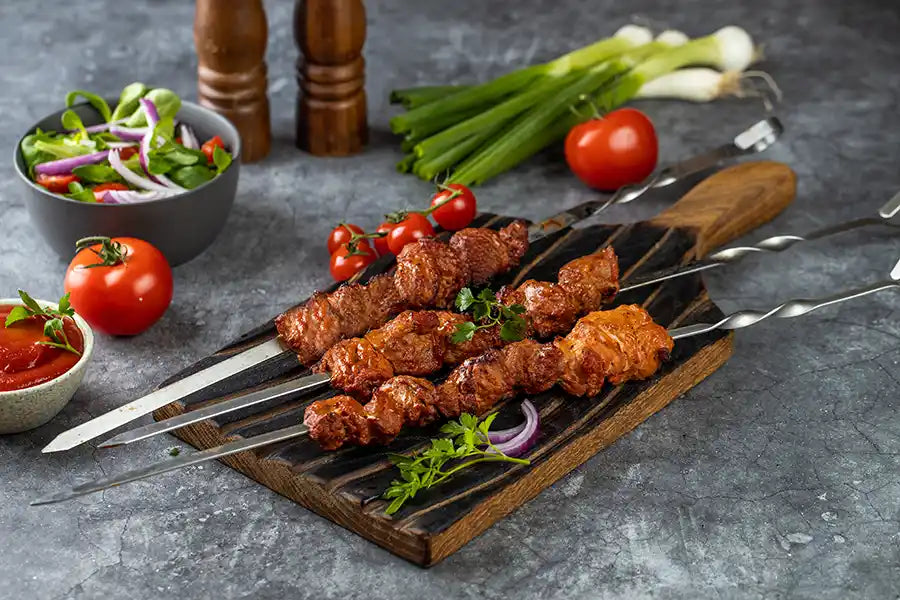 Schweine-Schaschlik Tomate angerichtet auf einem rustikalen Holzbrett mit roten Zwiebeln und Petersilie. Dekoriert mit Frühlingszwiebeln, Tomate, Sosse, Pfeffer- und Salzmühle aus Holz und Salat.