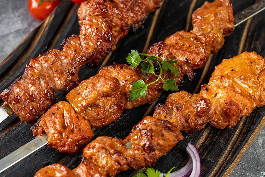 Schweine-Schaschlik Tomate angerichtet auf einem rustikalen Holzbrett mit roten Zwiebeln und Petersilie. Dekoriert mit Tomate.