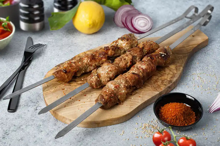 Schweine-Schaschlik Yakusa angerichtet auf einem Holzbrett. Garniert mit Sesam und Chili. Dekoriert mit Besteck, Zintrone, roten Zwiebeln, Cocktail-Tomaten und Sosse.